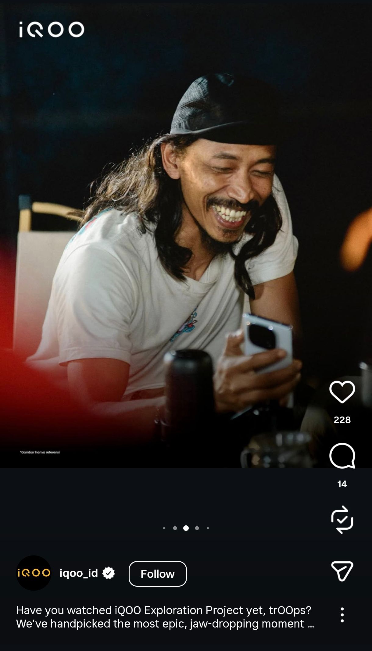 Person in white t-shirt smiling and looking at phone while sitting in dark room with ambient lighting.