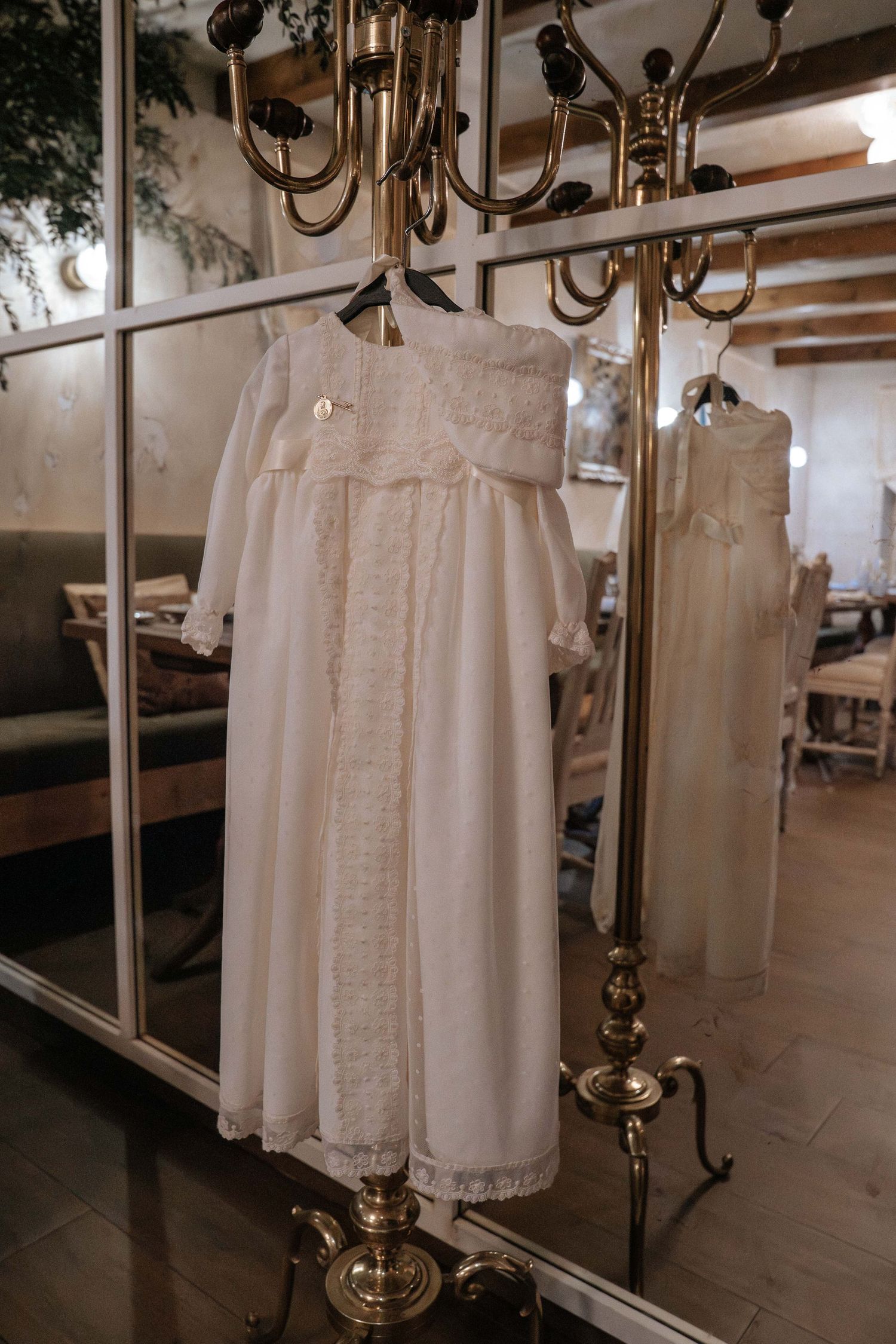 Vintage white christening gown with delicate lace details hangs elegantly against mirrored wall with brass chandelier reflection.
