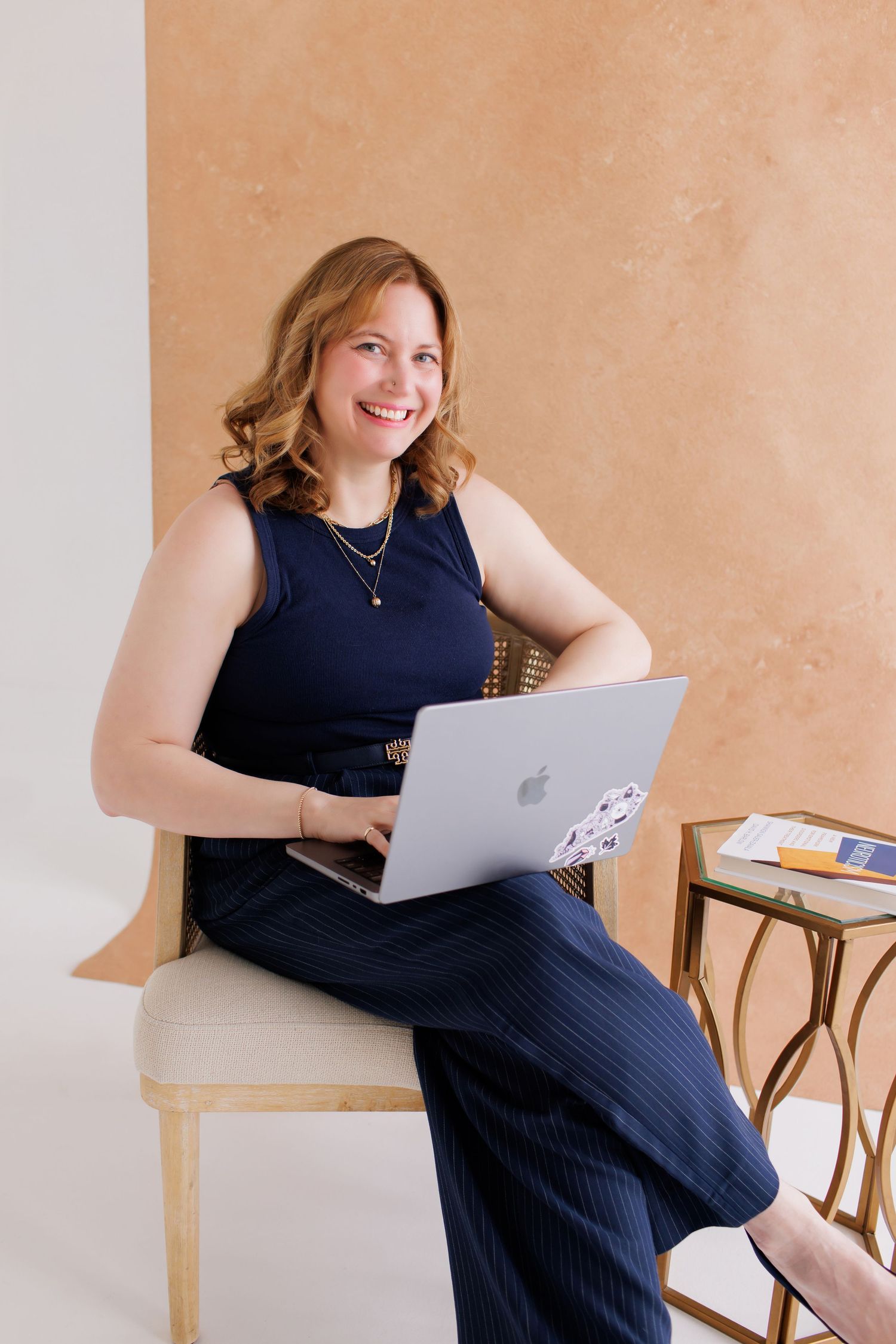 Professional wearing navy dress sits with MacBook laptop in modern minimalist setting with beige wall background.