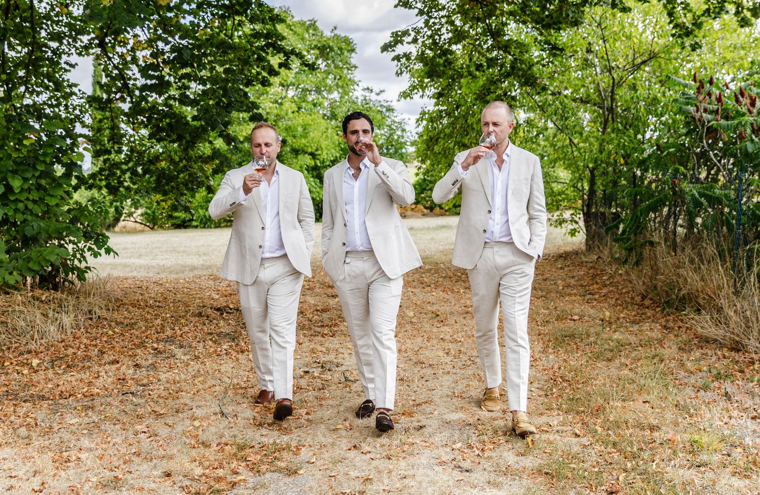 Three people in white outfits walk together through a lush garden pathway on a sunny day.