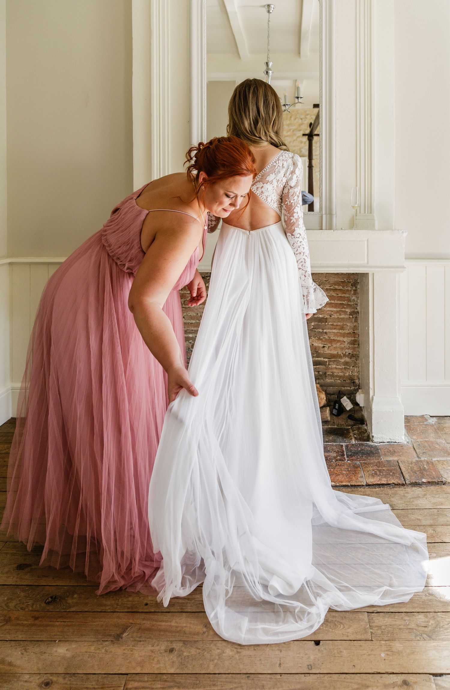A bridesmaid in a dusty rose gown helps adjust a flowing white wedding dress train in an elegant indoor setting.