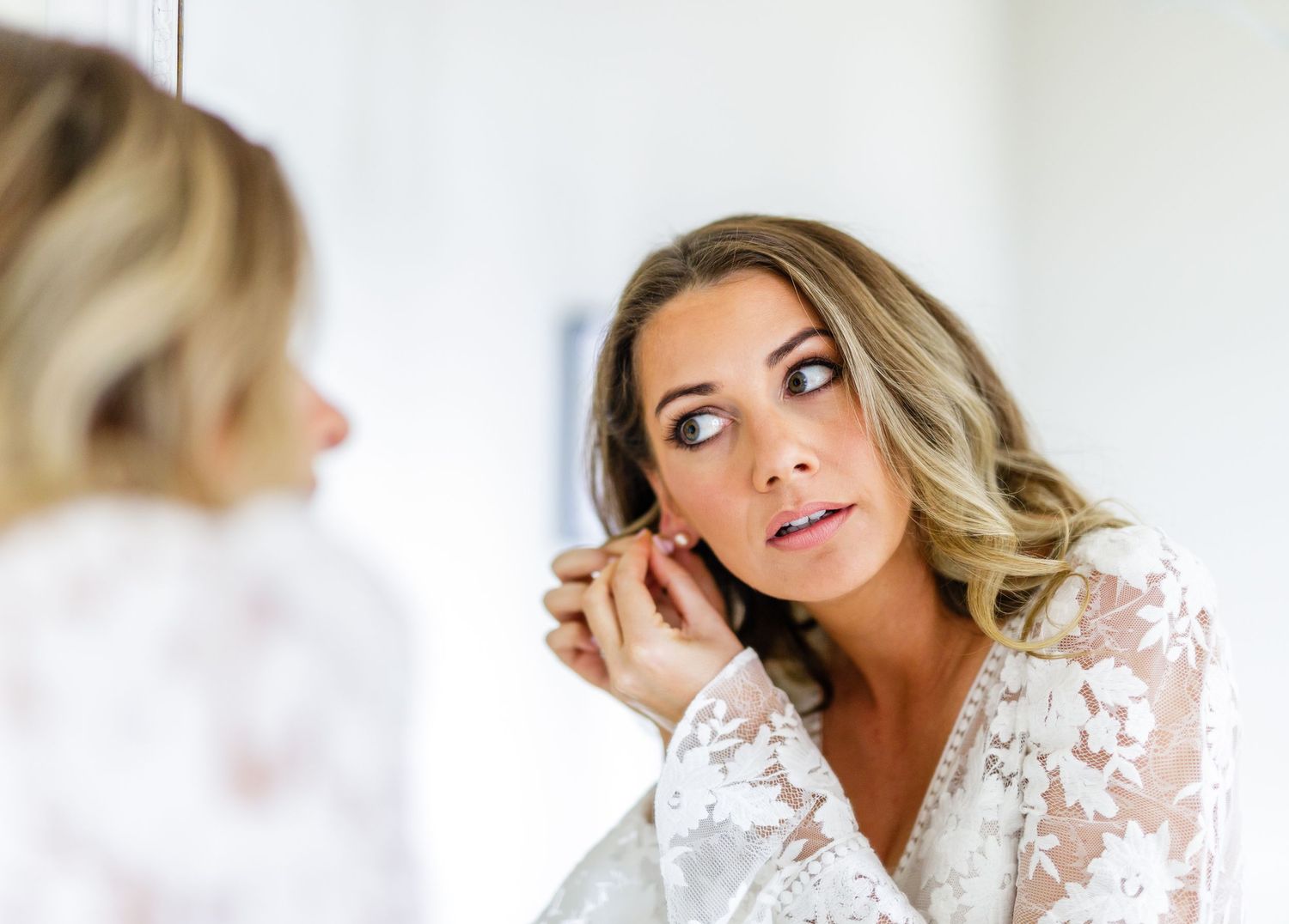 A bride in a white lace robe puts on earrings while looking in a mirror during wedding preparations.