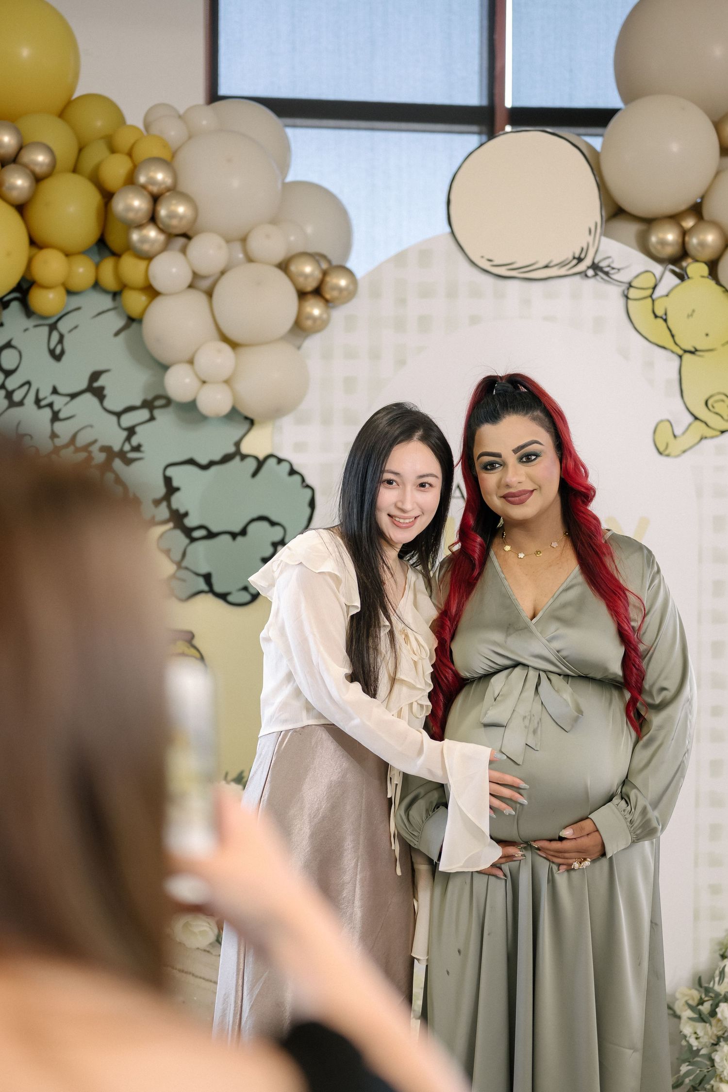 Two people pose together at a baby shower celebration with balloon decorations and pastel decor in the background.