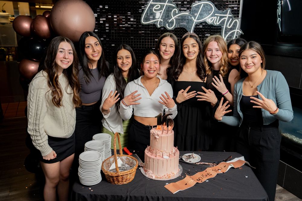 Group celebrating at a party with pink cake, wine glasses and festive decorations in a dimly lit venue.