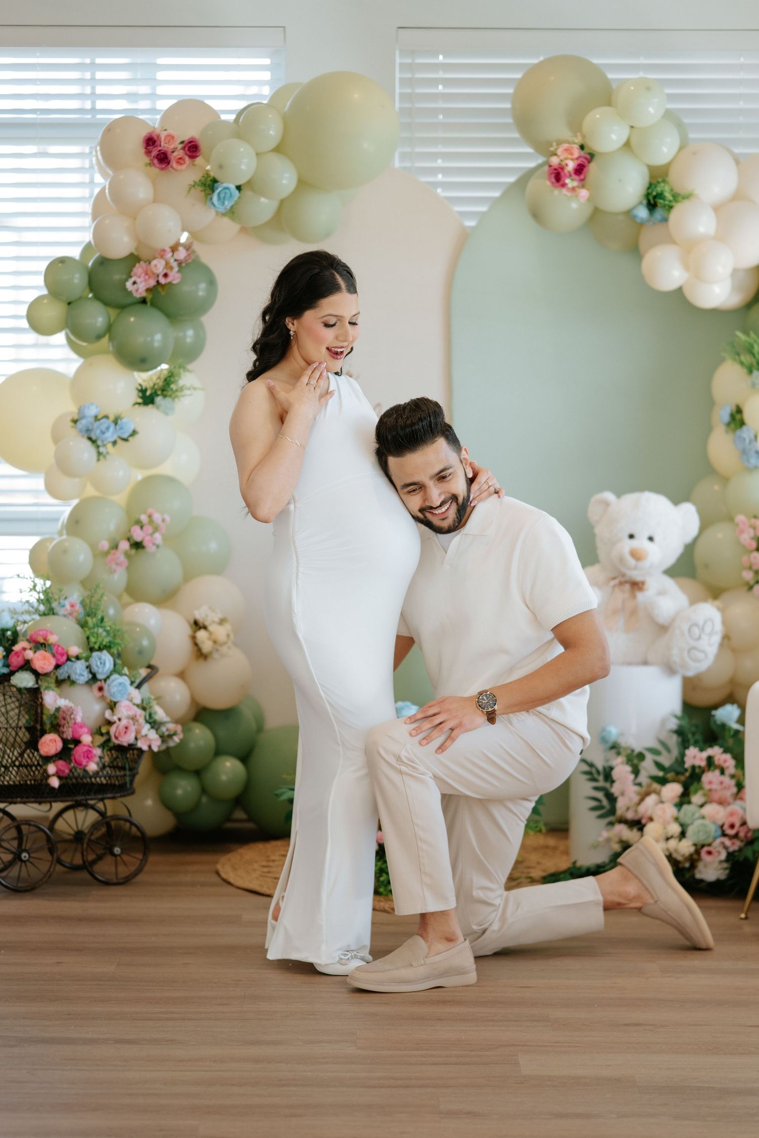 Couple at a baby shower with balloon arch decor, man kneeling beside pregnant woman in white dress.