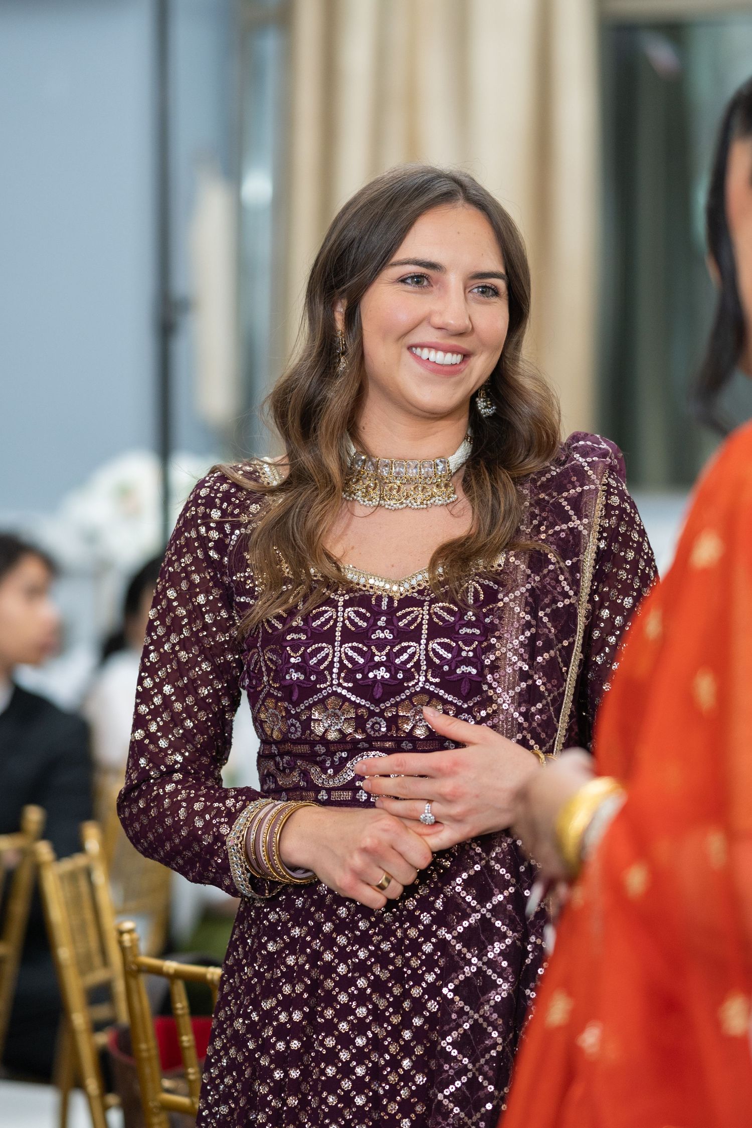 Woman in burgundy sequined dress smiles while seated at formal event with gold chairs visible in background.