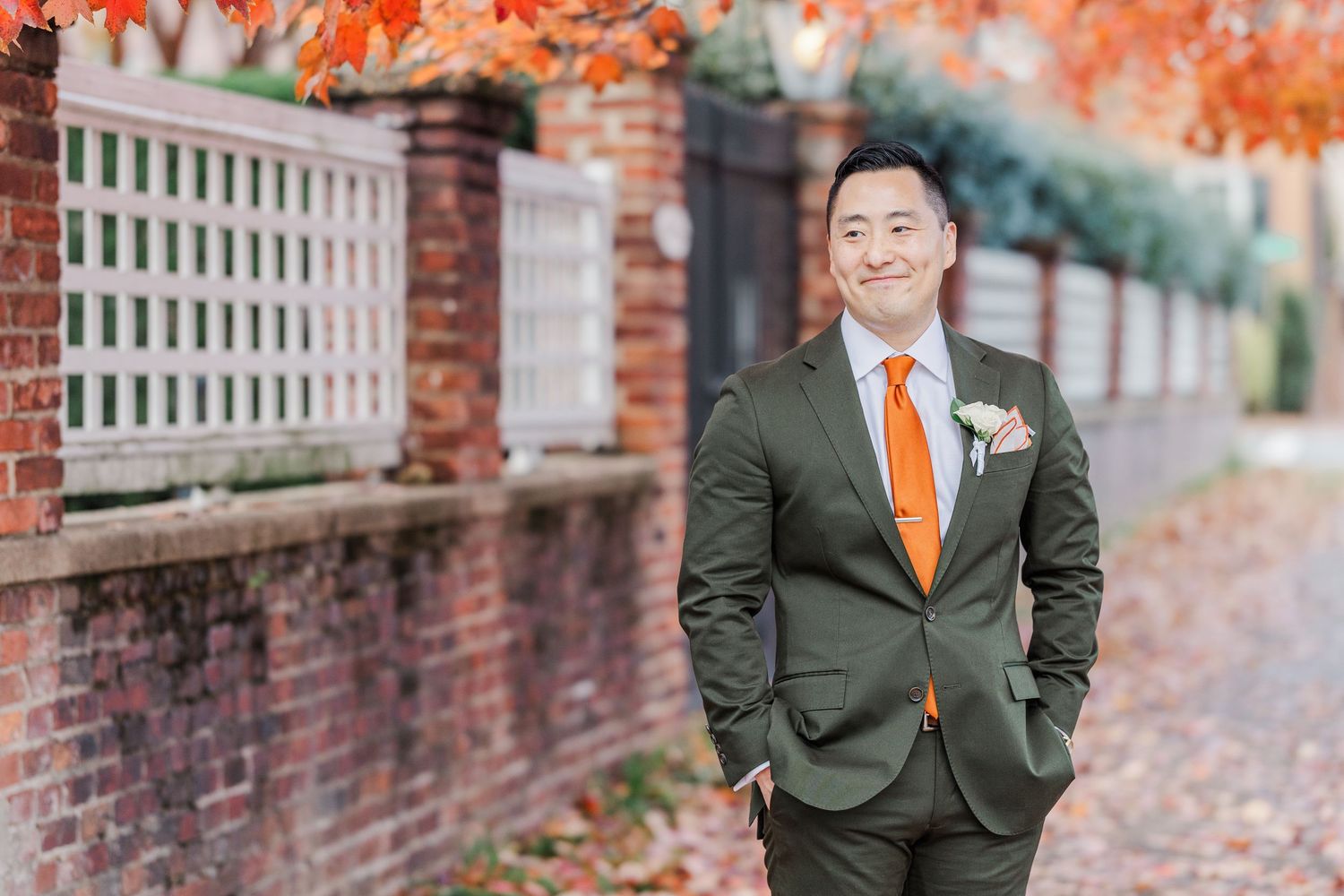 A person in a dark suit and orange tie stands against a brick wall with autumn leaves in the background.