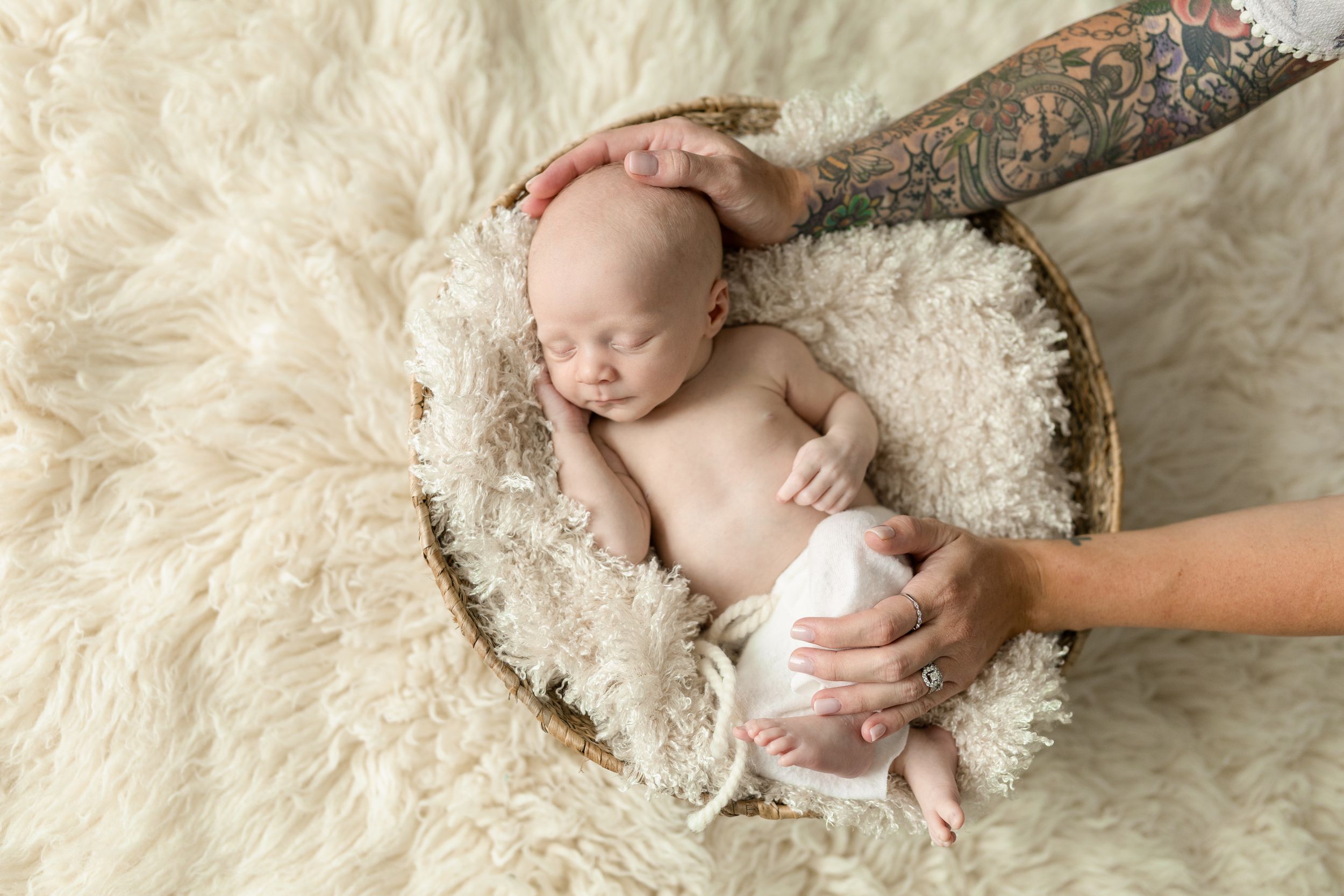 A newborn baby lies peacefully on soft white fur while tiny hands grasp a rustic wooden branch.