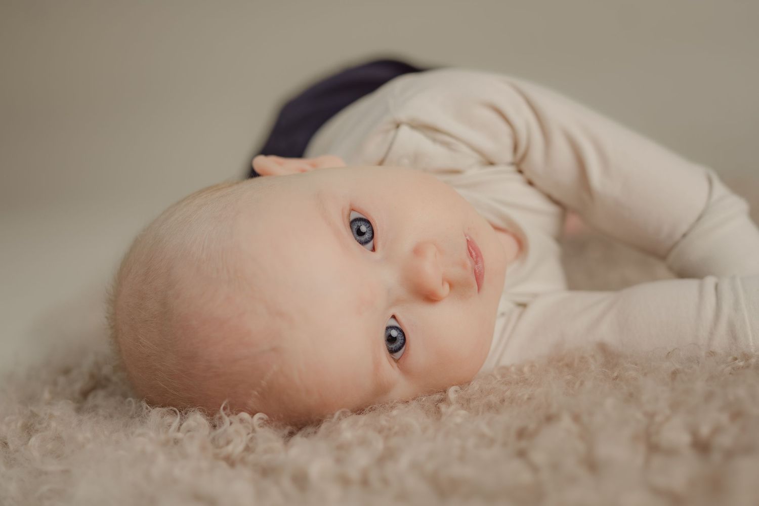 A baby lying on soft beige carpet gazes up with bright blue eyes while wearing a cream colored outfit.