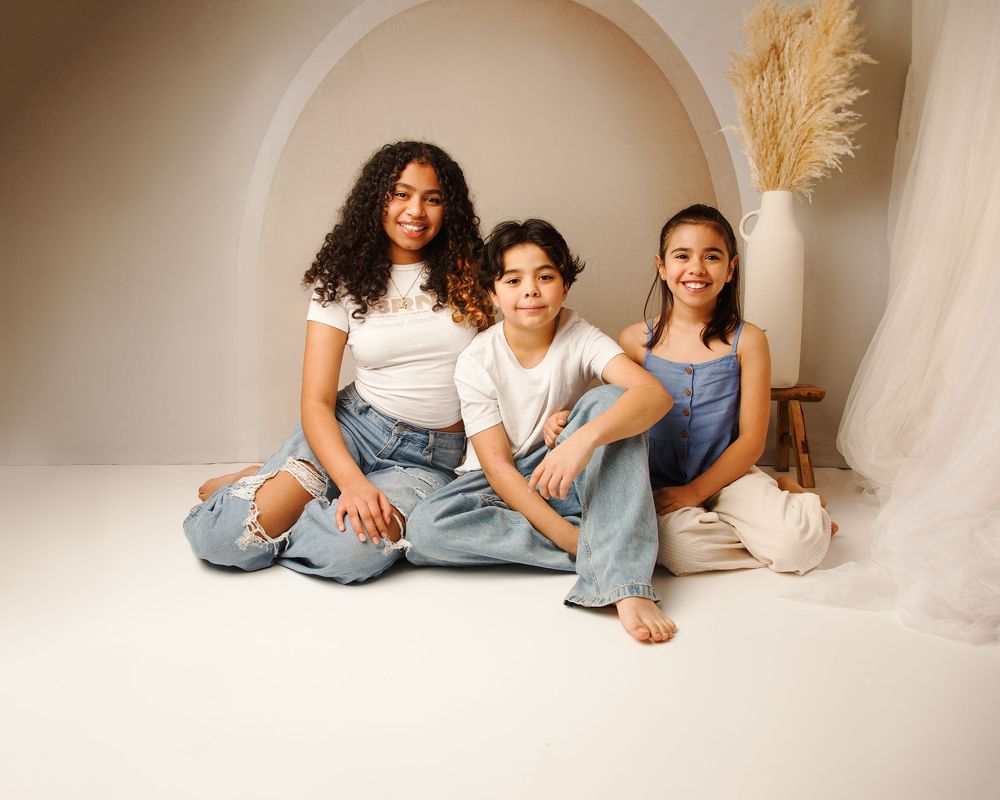Three people in casual denim and white outfits pose together in a minimalist studio setting with pampas grass decor.