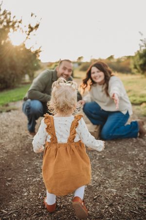 A toddler in brown overalls walks towards a couple sitting on the ground during golden hour sunset.