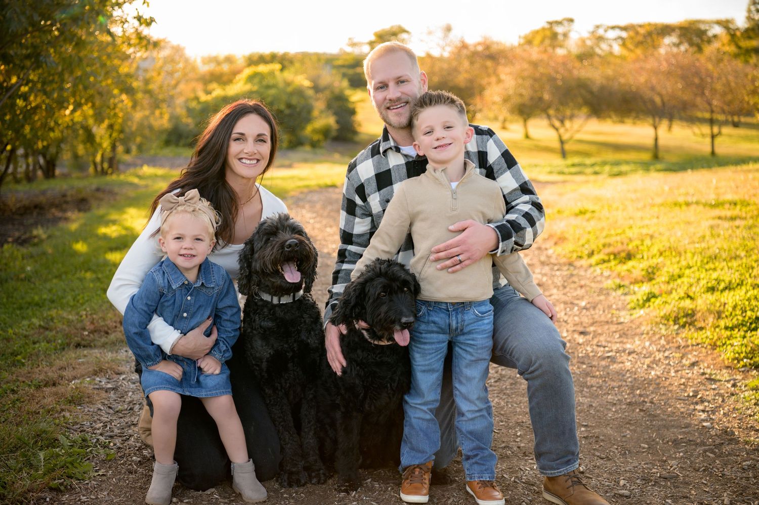 A family and their large black Newfoundland dog pose together on a dirt path during golden hour sunset.