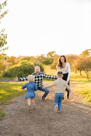 Children run playfully down a dirt path at sunset while an adult follows behind in a golden-lit rural setting.