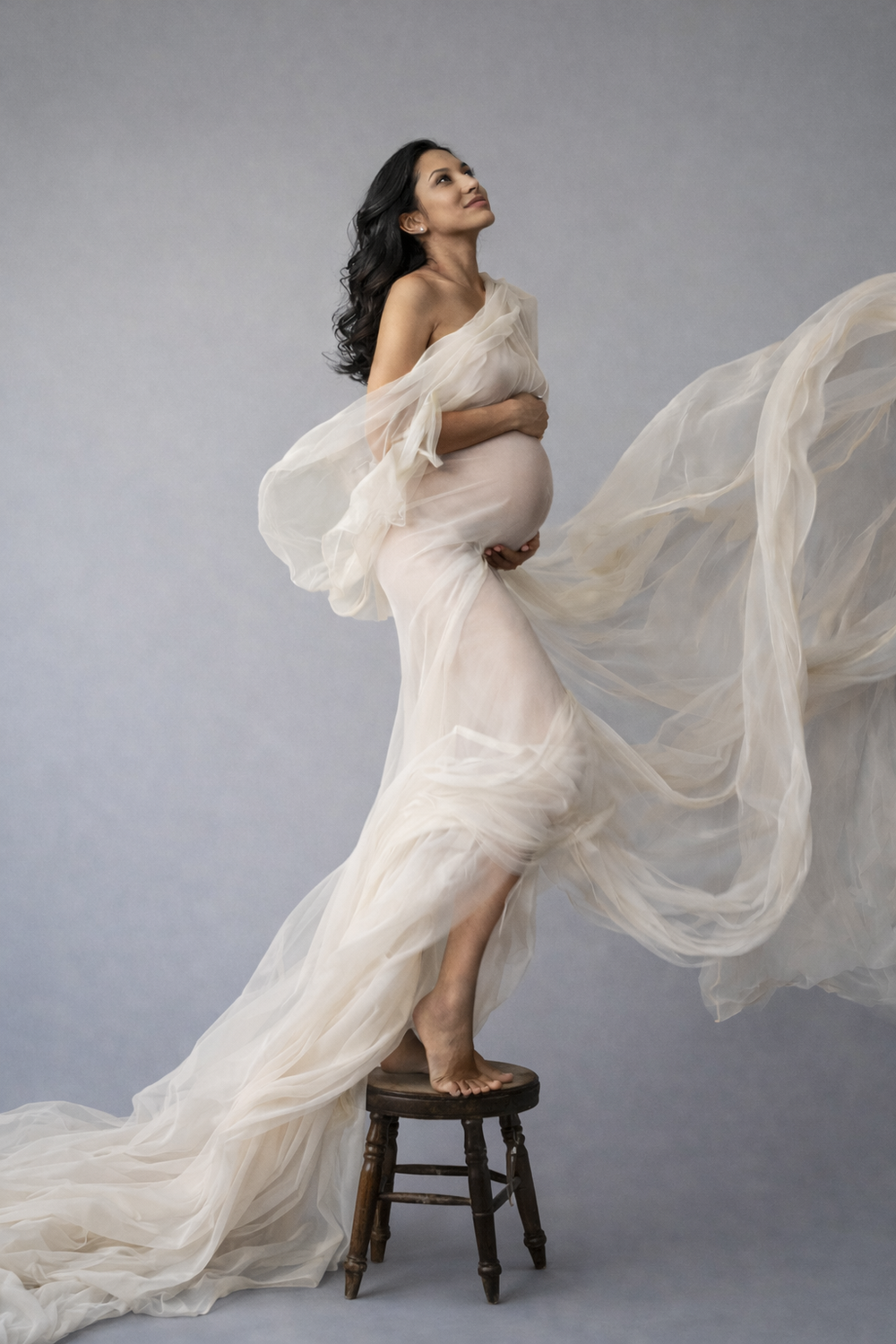 Pregnant woman in flowing ivory tulle gown poses gracefully on wooden stool against gray studio background.