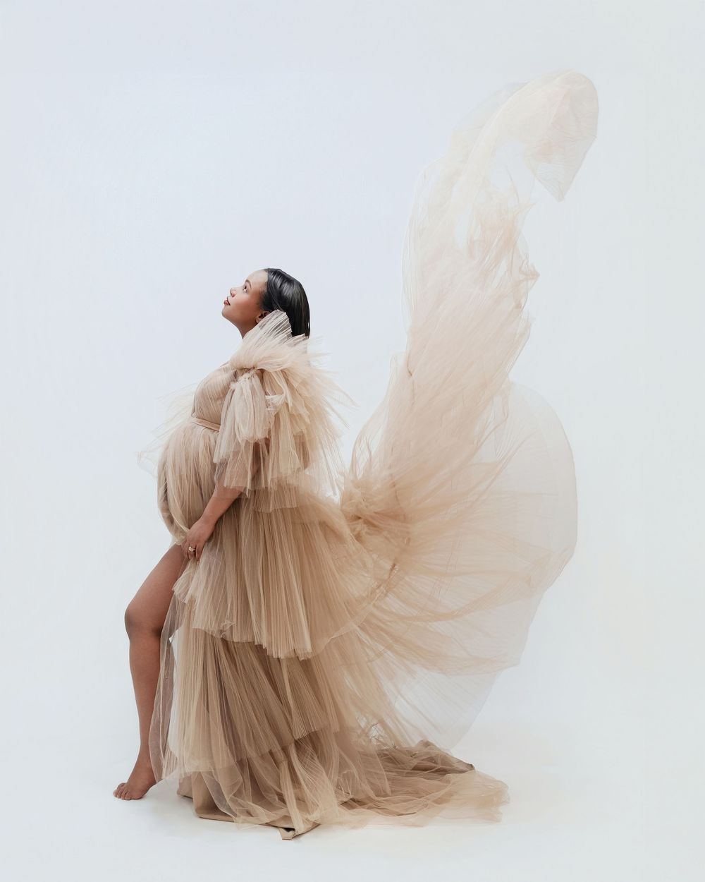 Pregnant woman in flowing champagne tulle gown poses gracefully against white background with fabric billowing dramatically.