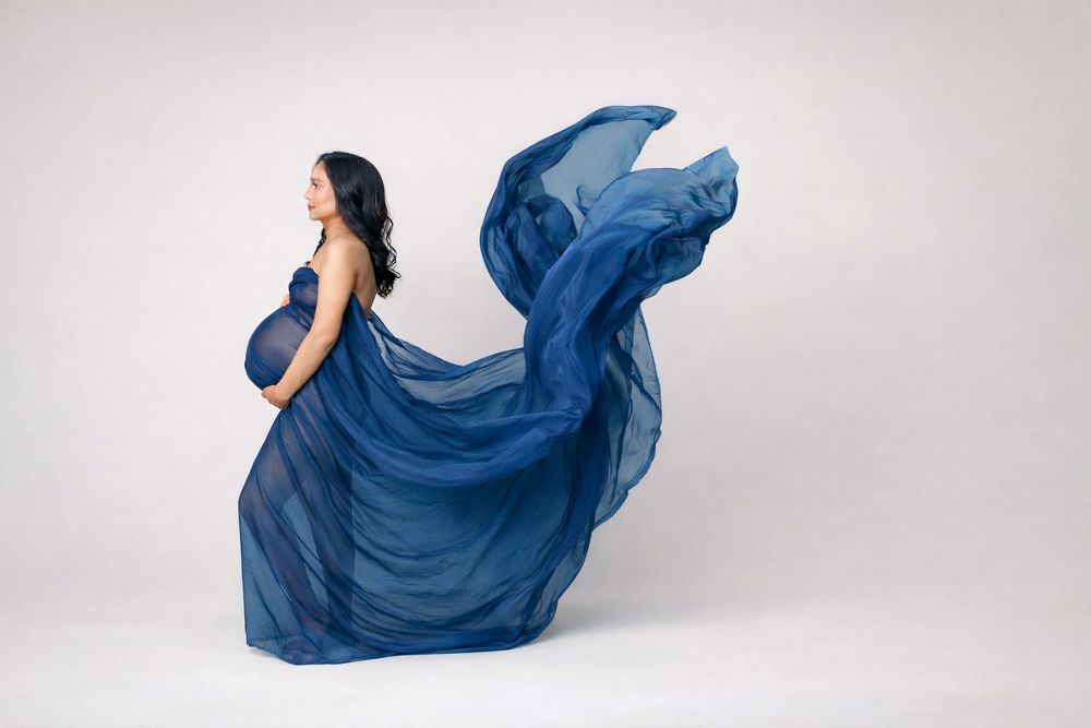 Pregnant woman in flowing blue chiffon gown with fabric billowing dramatically against white background.
