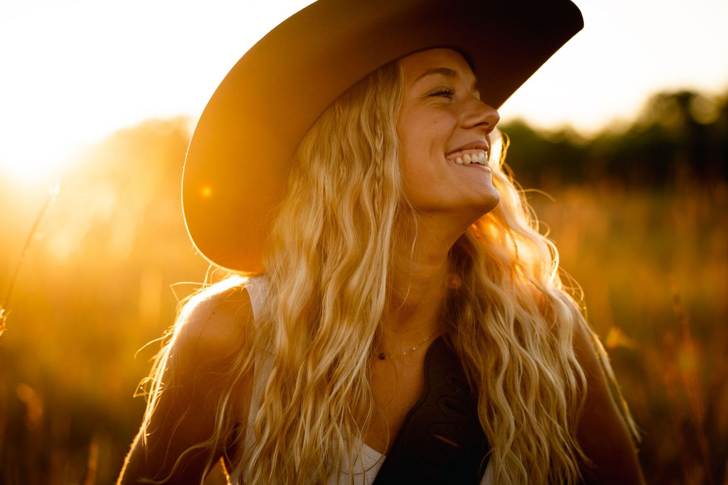 A woman in a white tank top laughs joyfully against a sunset sky with a rainbow in the background.