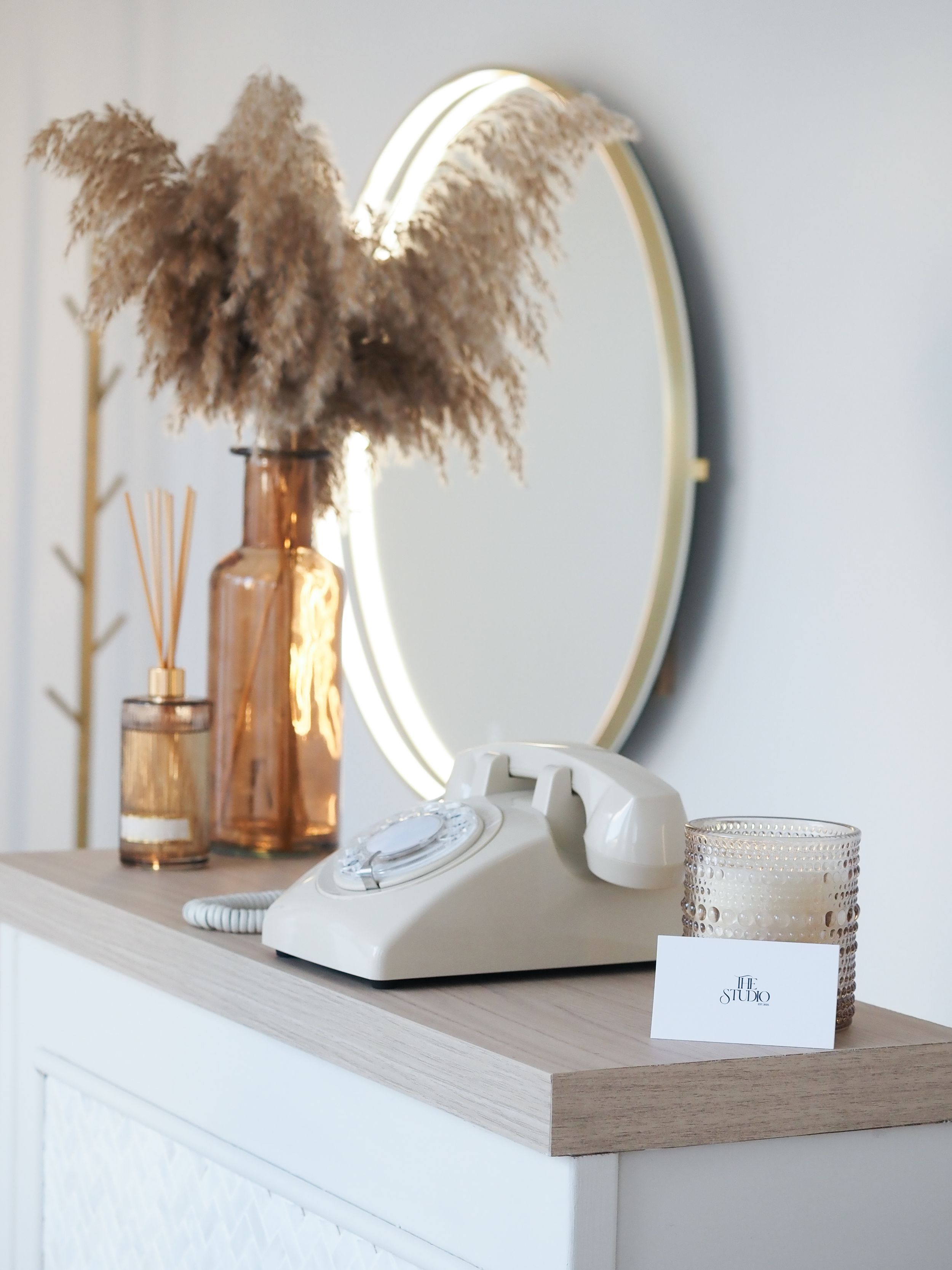 Modern minimalist home decor with round mirror, dried pampas grass, and wooden shelf against light gray wall.