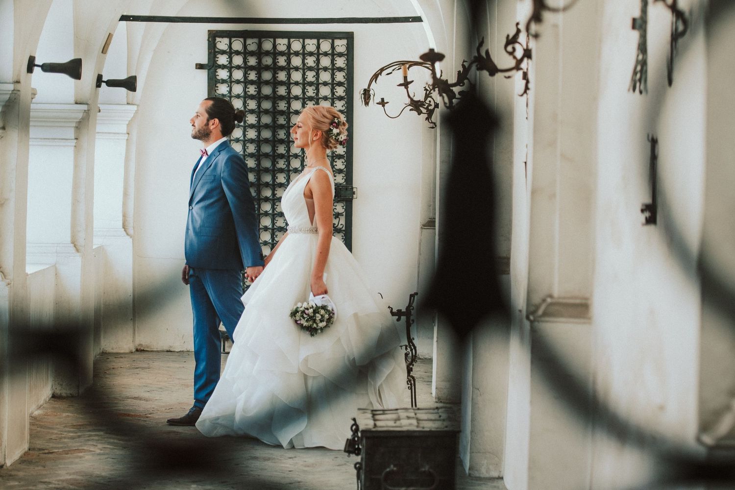 Artistic wedding photo of couple in formal attire standing in an elegant corridor with architectural details.