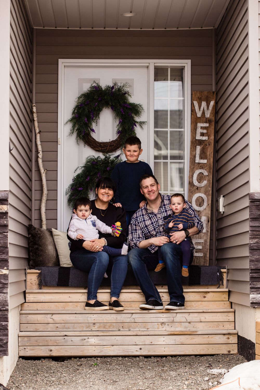 People sit casually on wooden porch steps with decorative welcome letters and evergreen wreaths.
