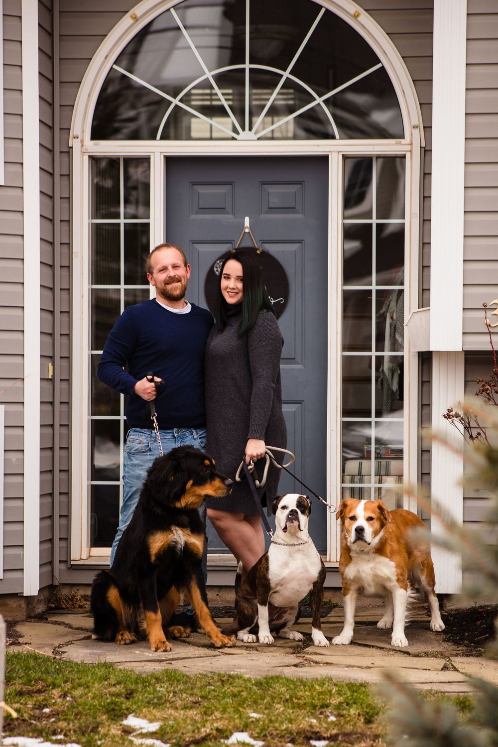 Several dogs of different breeds pose with their owners on front steps of gray house.