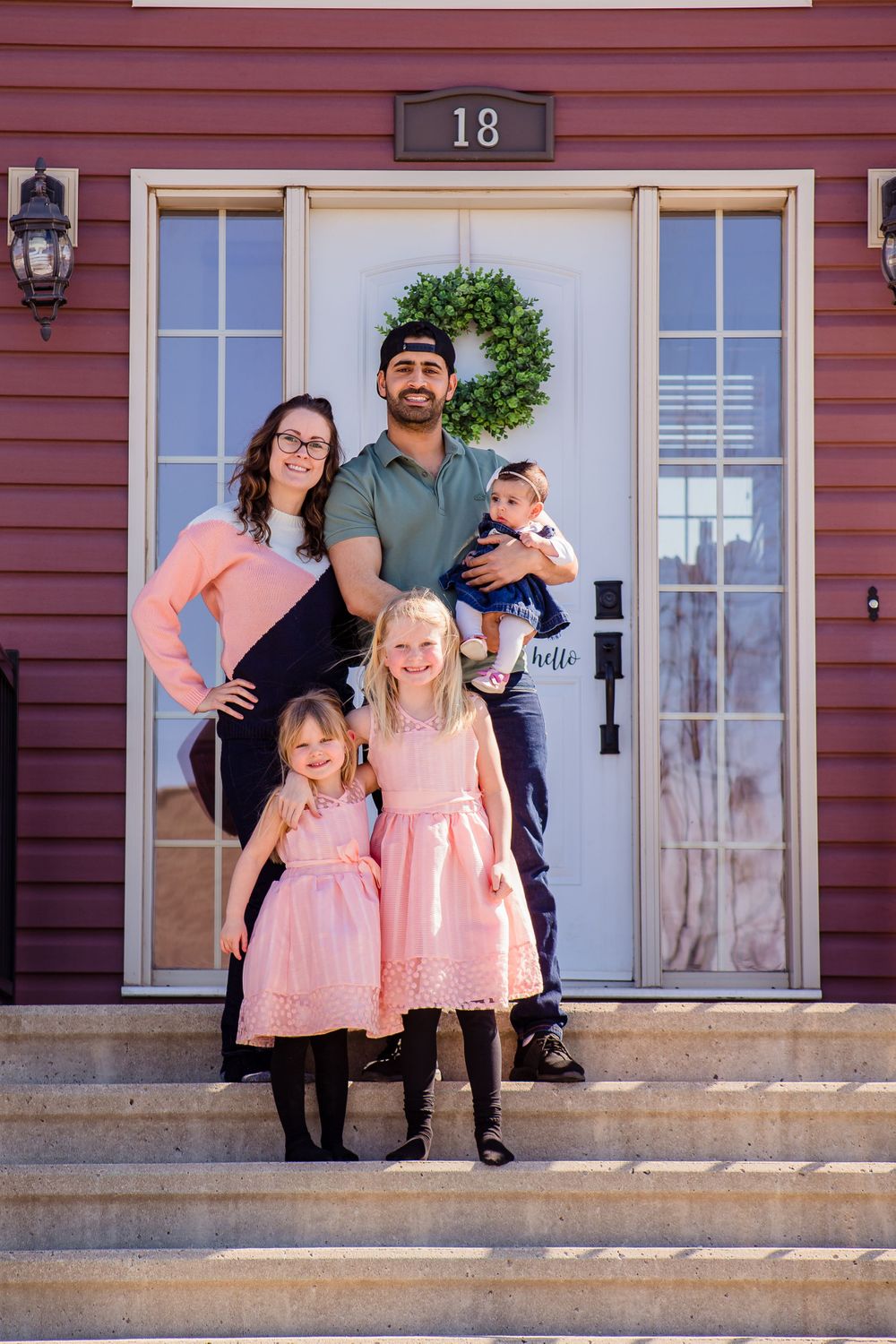 People posed on brick steps of a red house with white trim and house number 18.