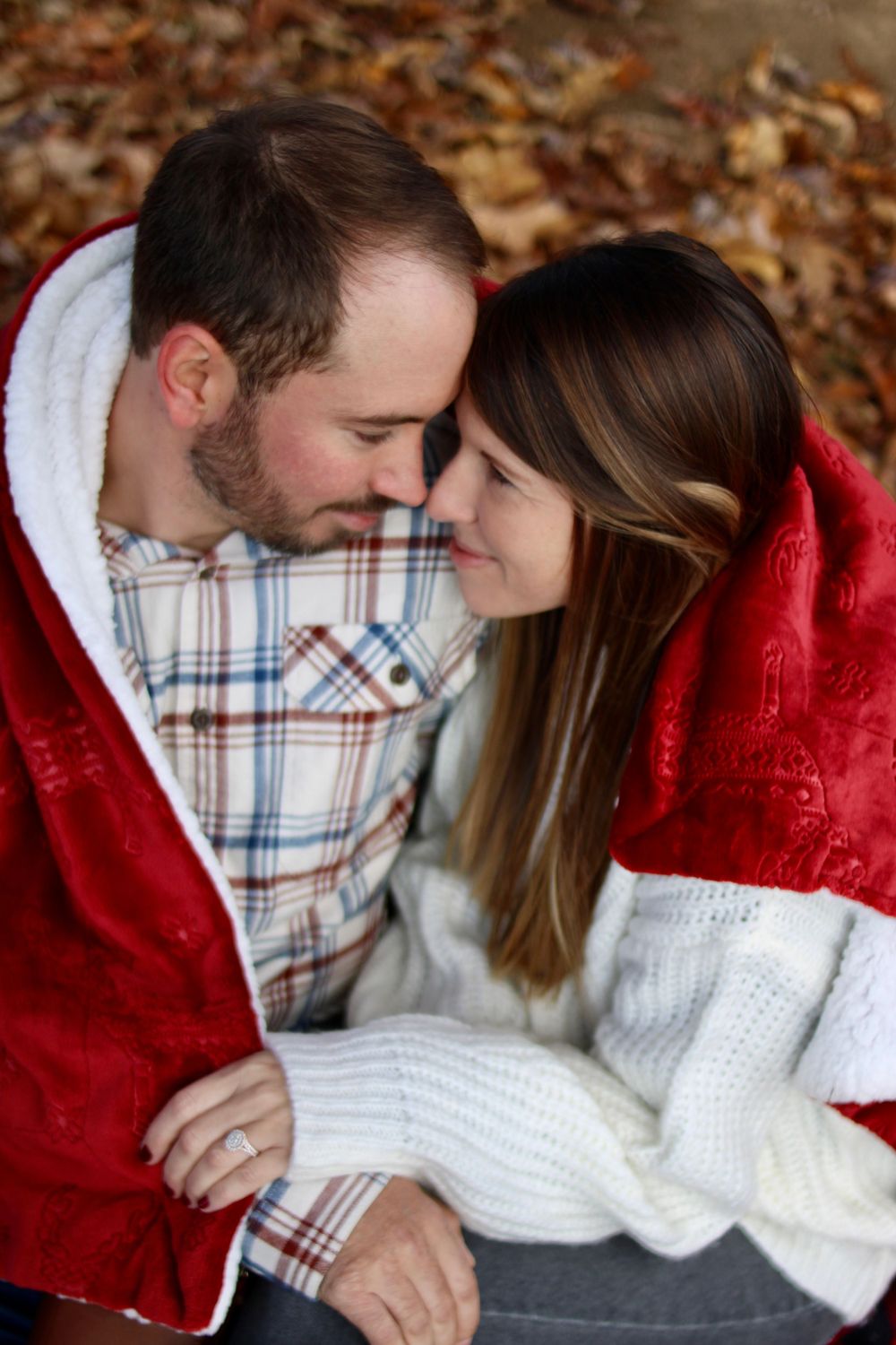 Couple cuddling under red blanket wearing plaid shirt and white sweater.