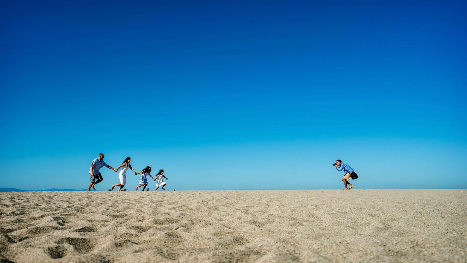 Silhouettes of people running on sandy dunes against bright blue sky.
