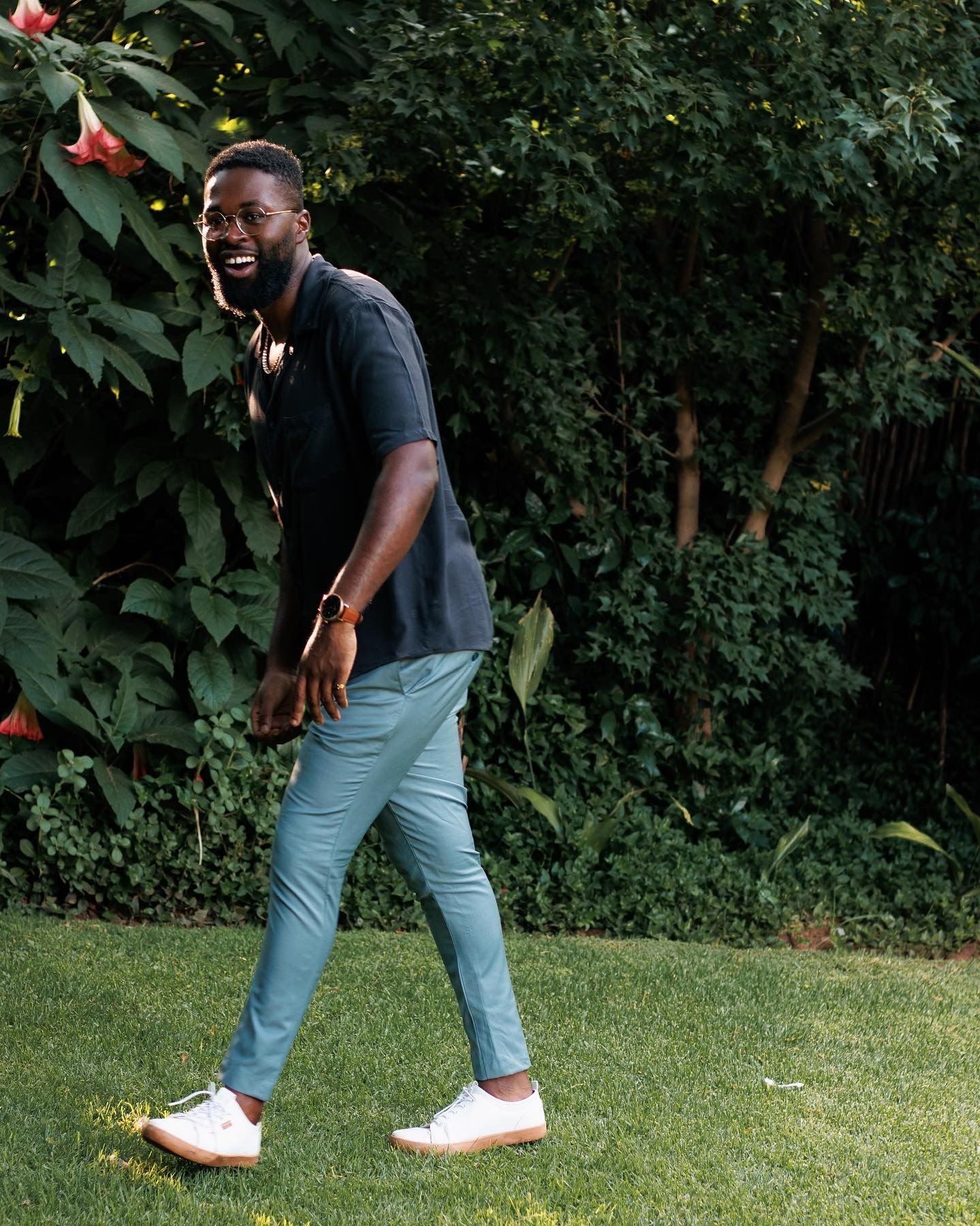 Person in casual attire strikes playful pose in garden setting with green foliage.