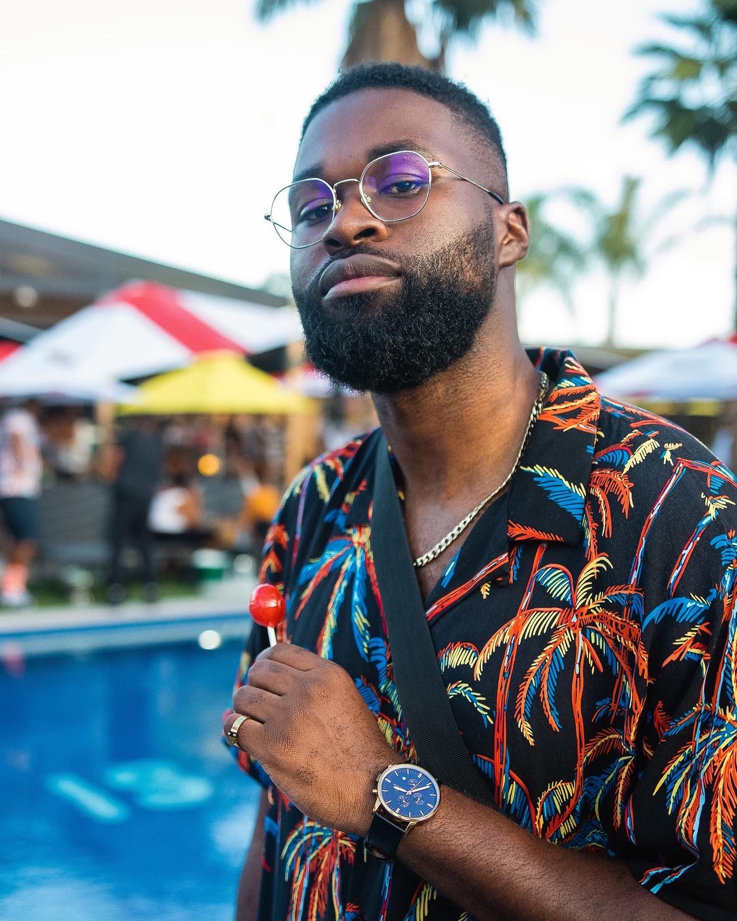 Person in tropical print shirt poses by pool wearing glasses and watch with palm trees in background.