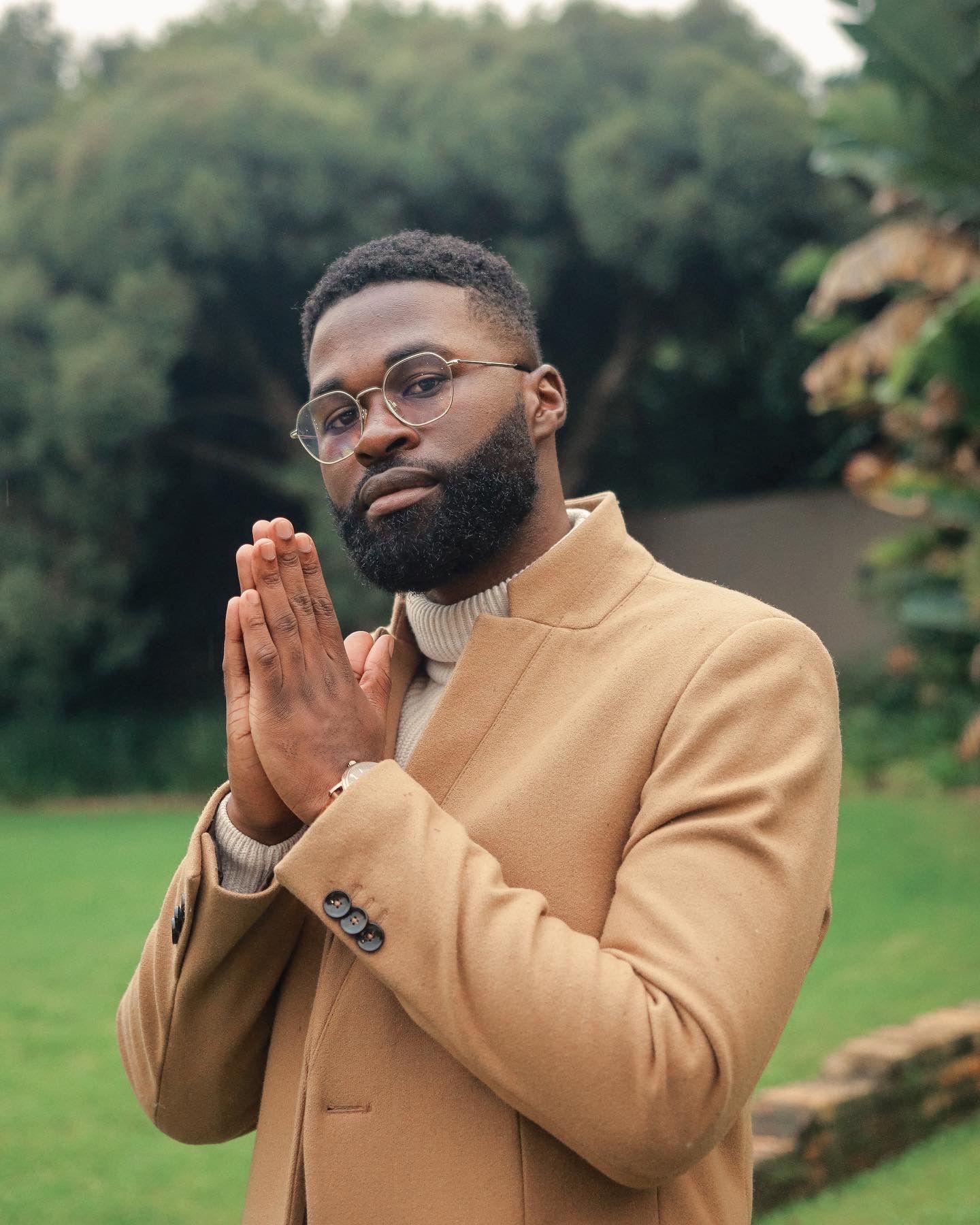 Person in a camel coat and glasses makes a prayer gesture outdoors in a garden setting.