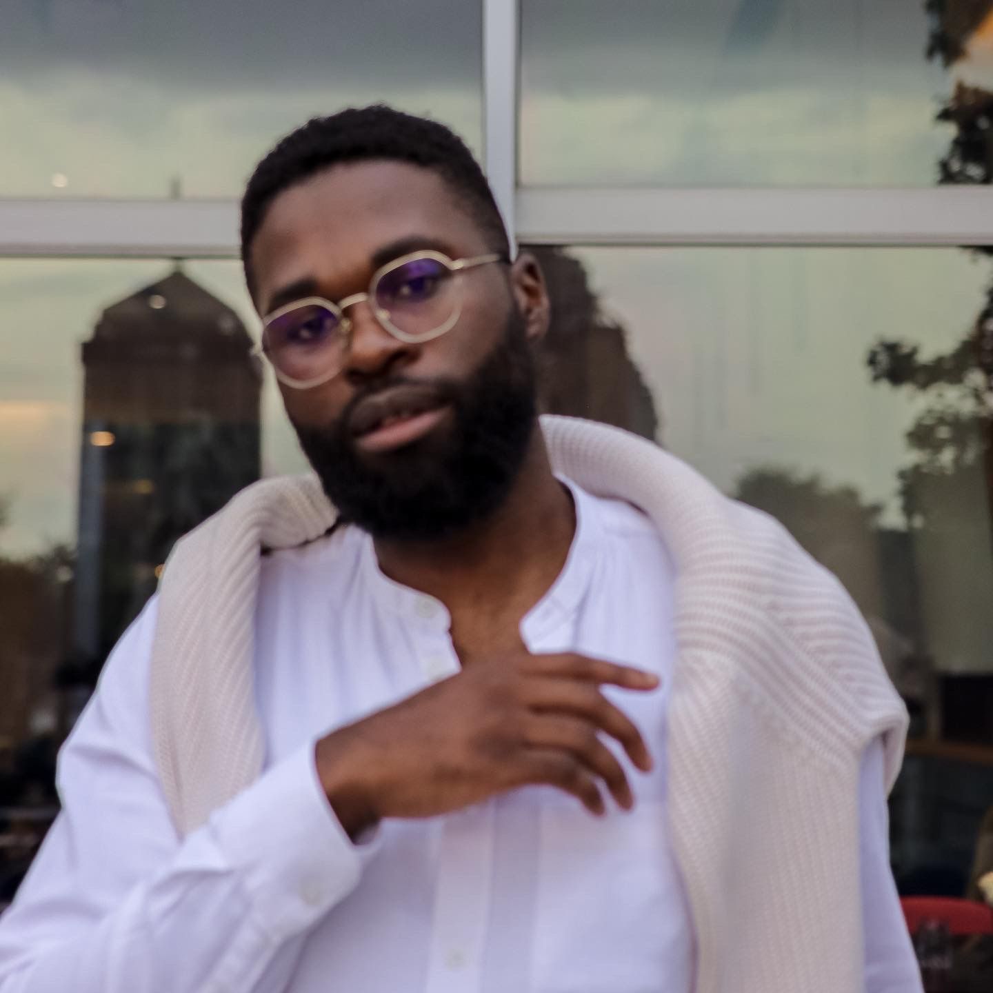 Person in a white shirt and cardigan poses casually outdoors wearing purple glasses.