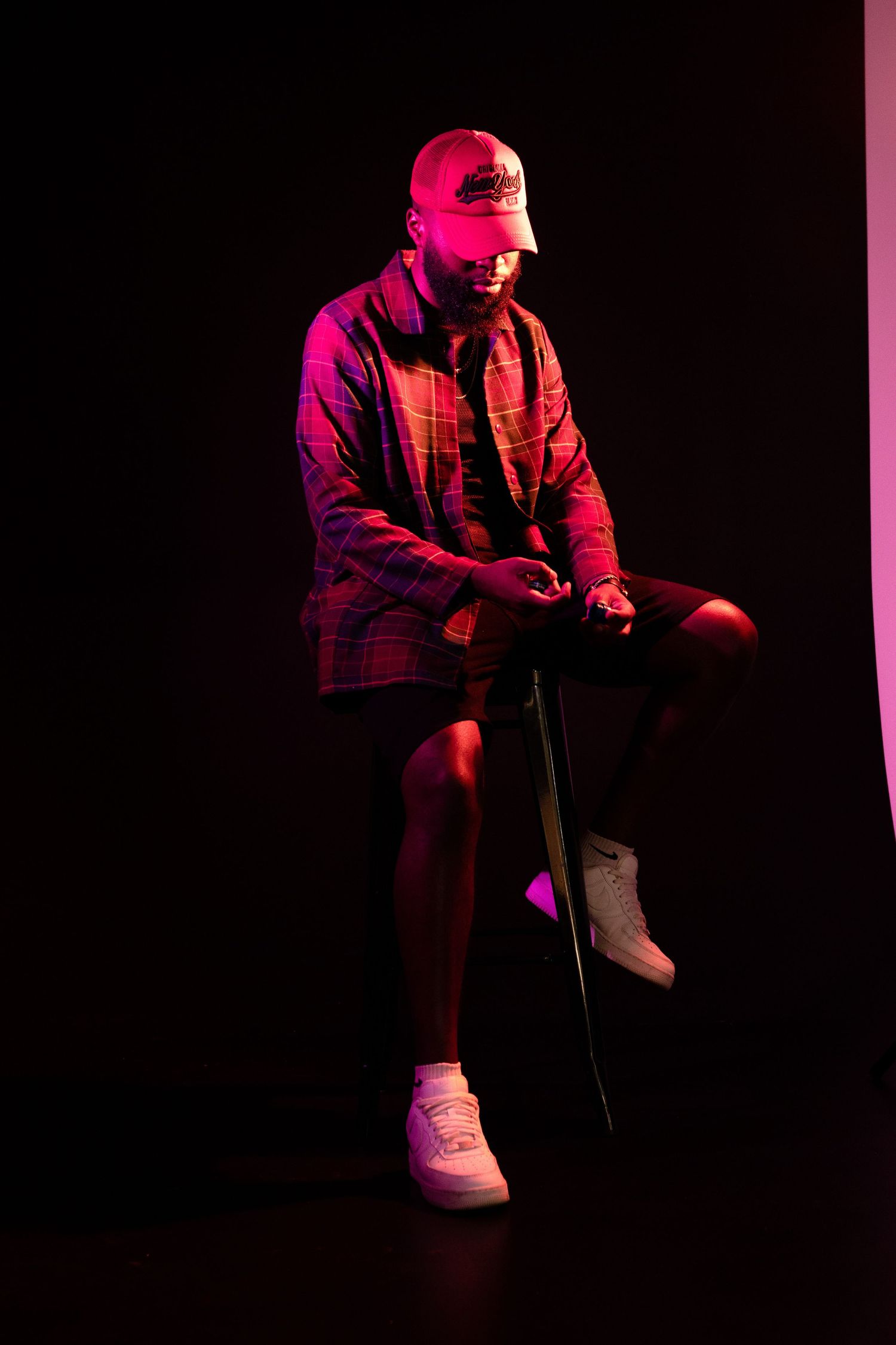 Figure poses on stool in red-lit studio wearing baseball cap, plaid shirt and white sneakers.