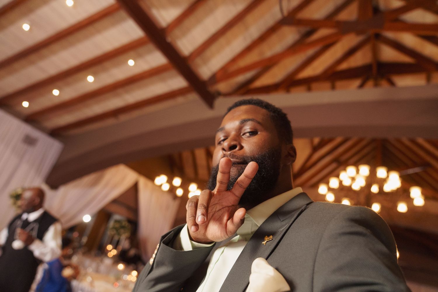 Person in a grey suit shows excitement with a peace sign gesture at an indoor wedding reception venue.