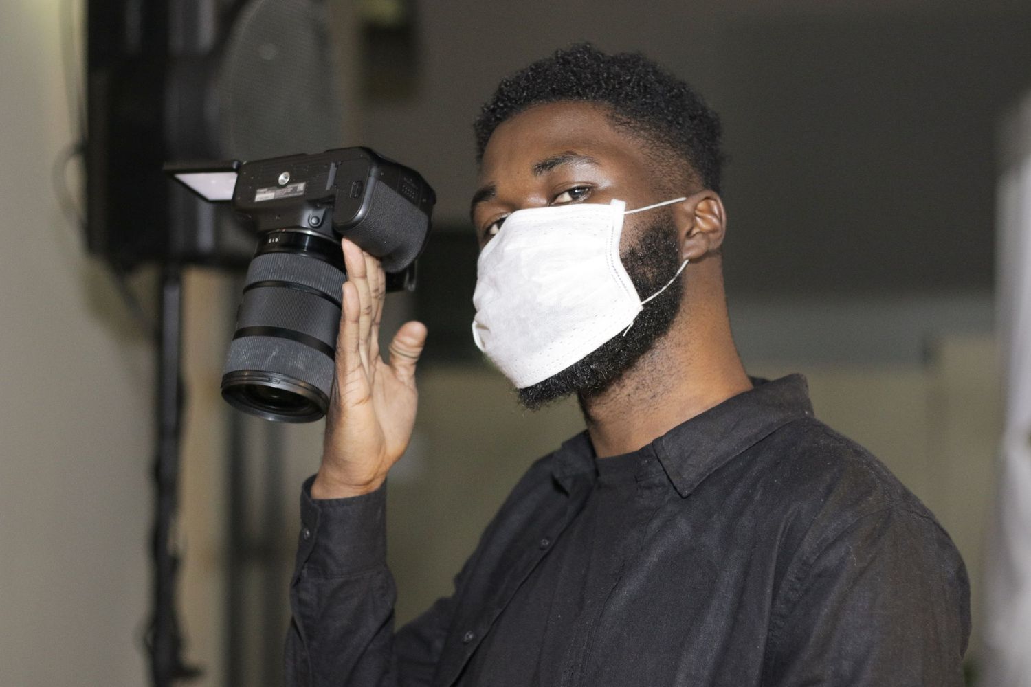 Photographer wearing a protective face mask holds up a professional camera in an indoor setting.