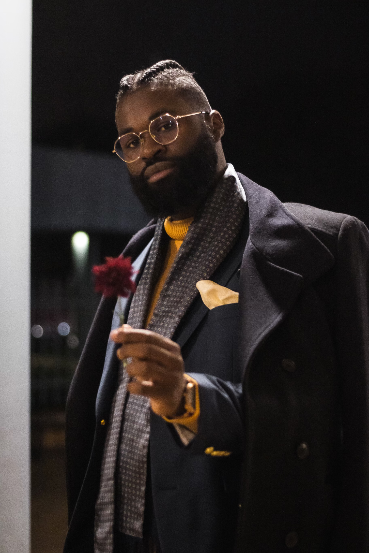 Person in formal attire with glasses and patterned scarf poses against dark background.