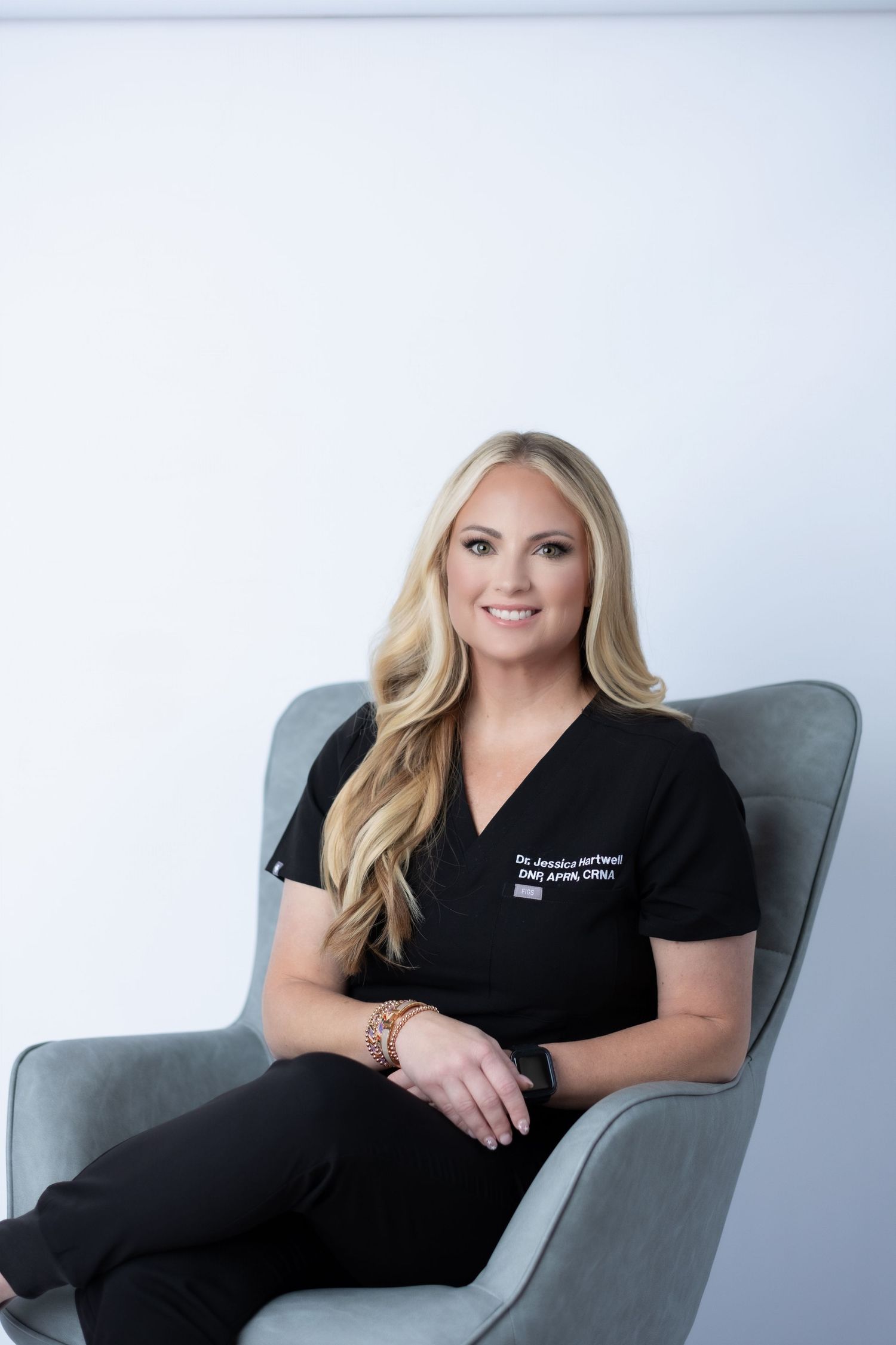Healthcare professional in black scrubs sits confidently in a modern grey chair against white background.