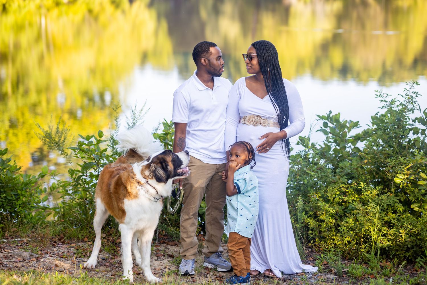 Family poses with their Saint Bernard dog by lake during maternity photoshoot.