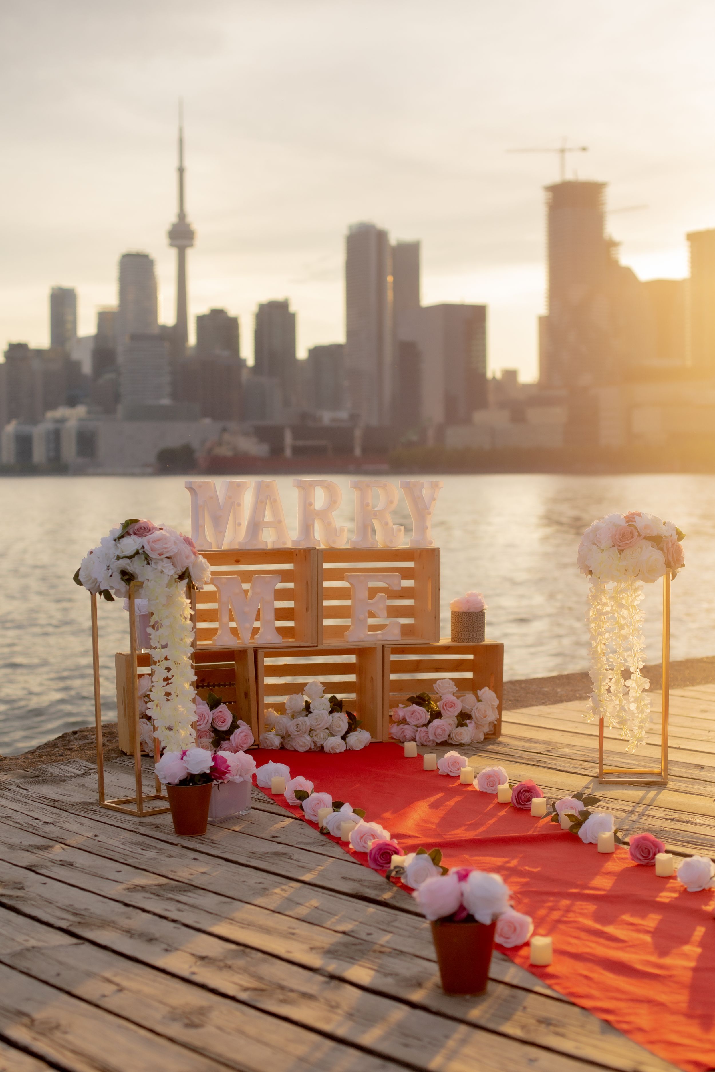 Romantic proposal setup with red carpet and floral decorations on a pier overlooking Toronto skyline at sunset.