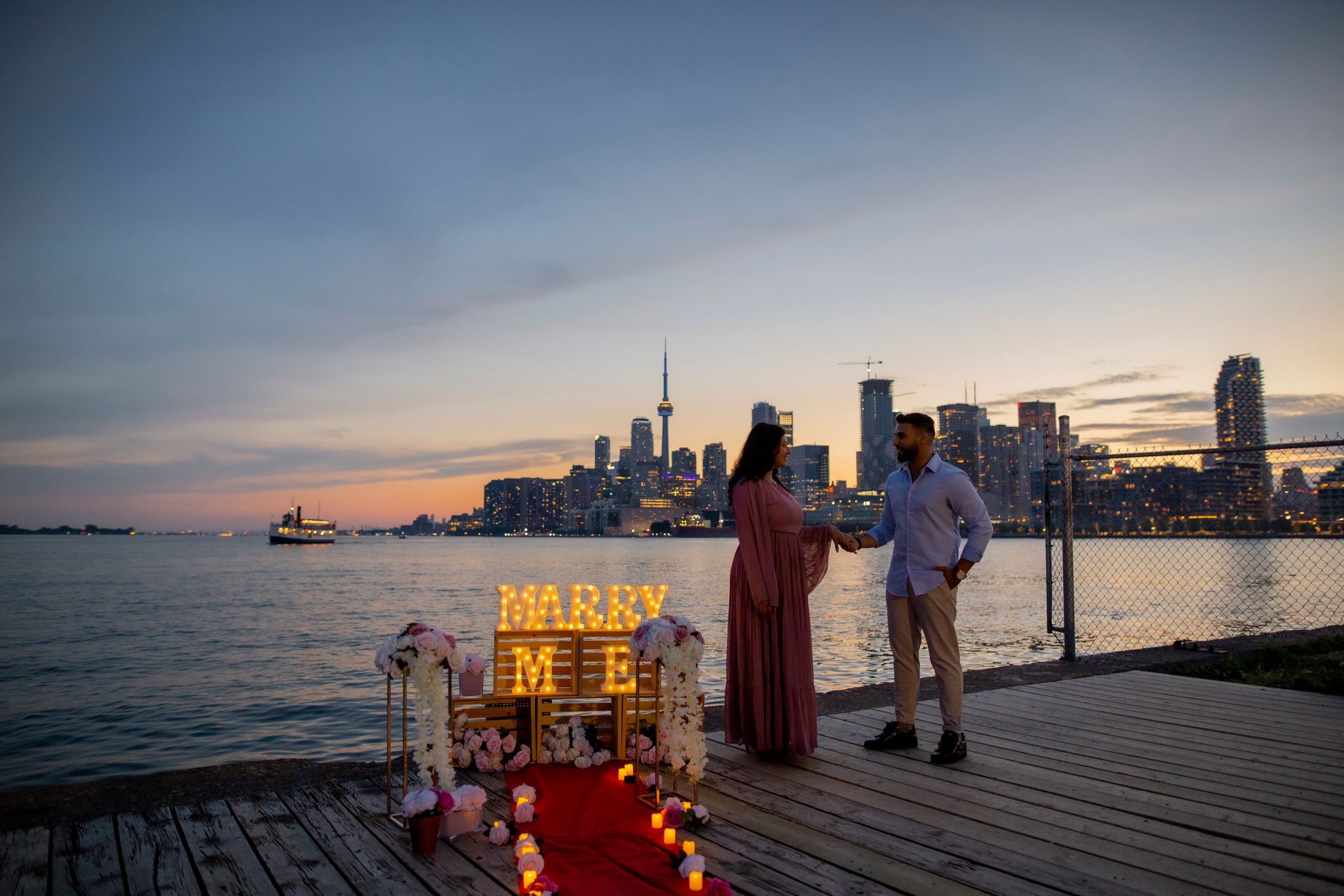Illuminated 'MARRY ME' sign on wooden pier with Toronto skyline at dusk.