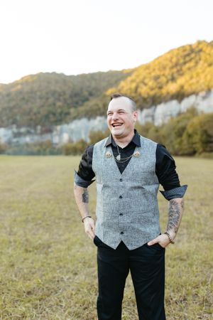 A person wearing a grey vest and black shirt smiles while standing in a field with mountains behind.