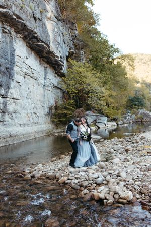 A couple in wedding attire poses by dramatic limestone cliffs and river rocks.