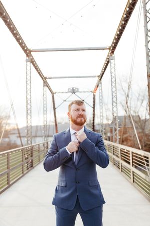 A groom in navy suit adjusts his tie while posing on an industrial metal bridge.