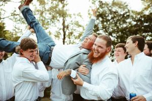 Groomsmen in white shirts playfully lifting and celebrating at outdoor wedding reception.