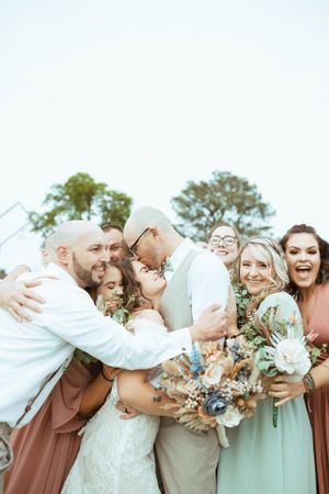 A joyful wedding party embraces and laughs together while holding floral bouquets.