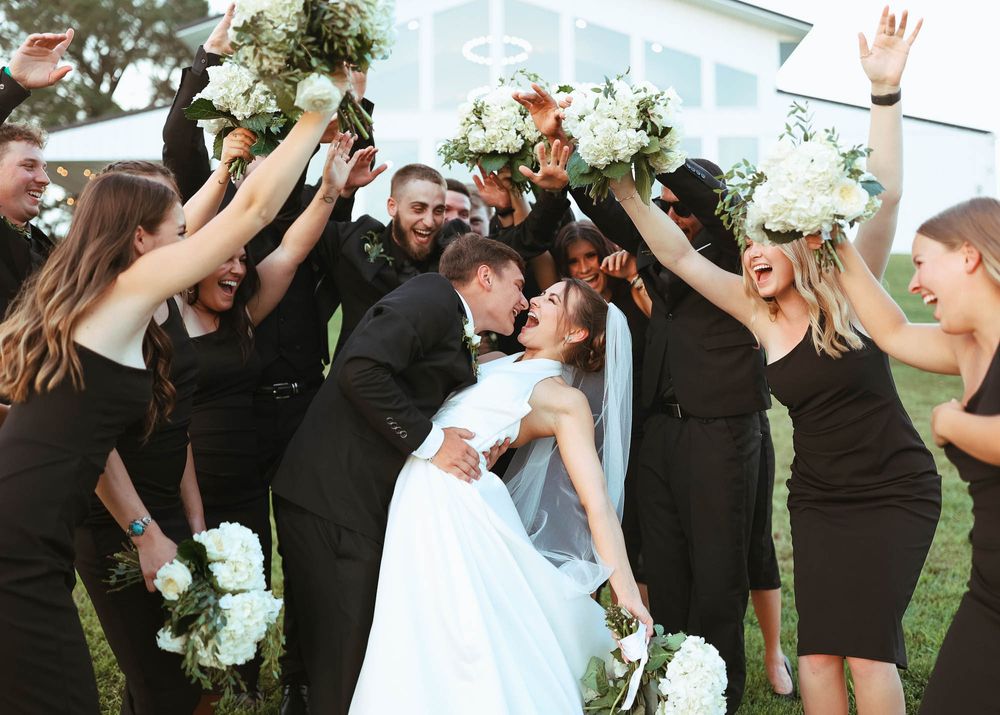 Wedding party holds bouquets overhead in celebration as couple shares romantic moment in center.