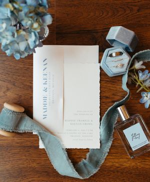 Wedding details flatlay with invitation, perfume bottle, ring box and blue silk ribbon.