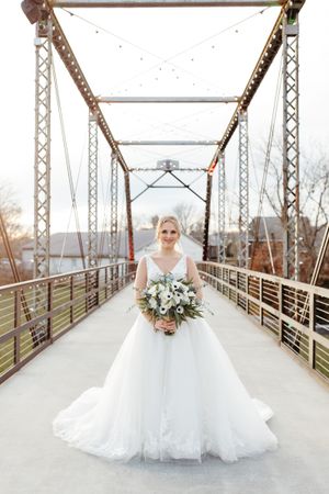 A bride in a white ballgown wedding dress stands on a metal bridge holding a floral bouquet at sunset.