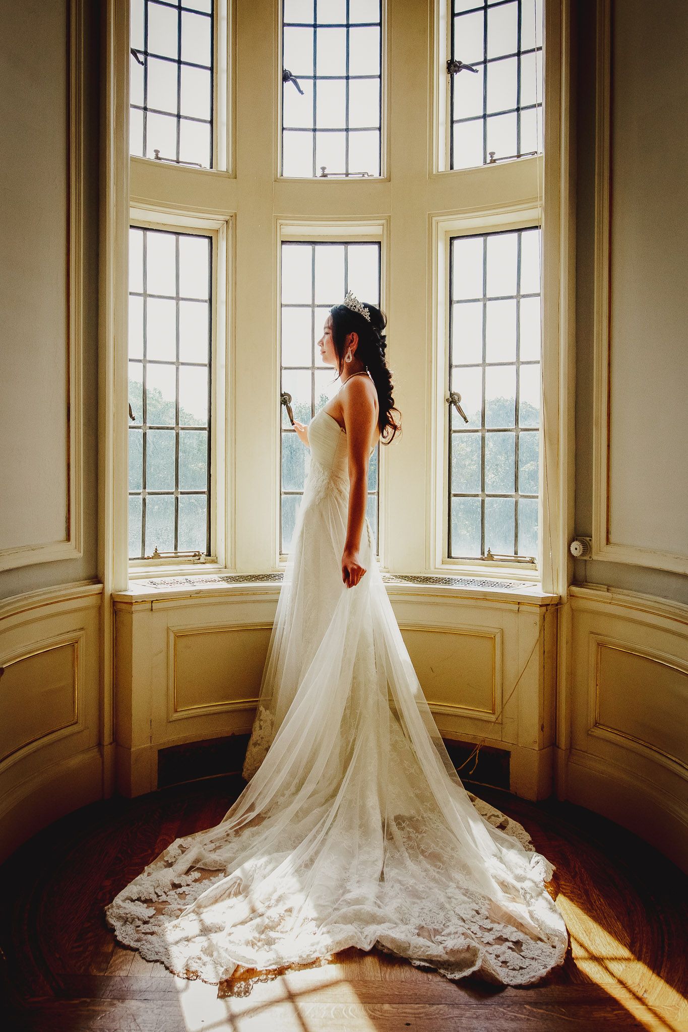 A bride in a fitted lace gown with long train stands before tall bay windows in golden sunlight.