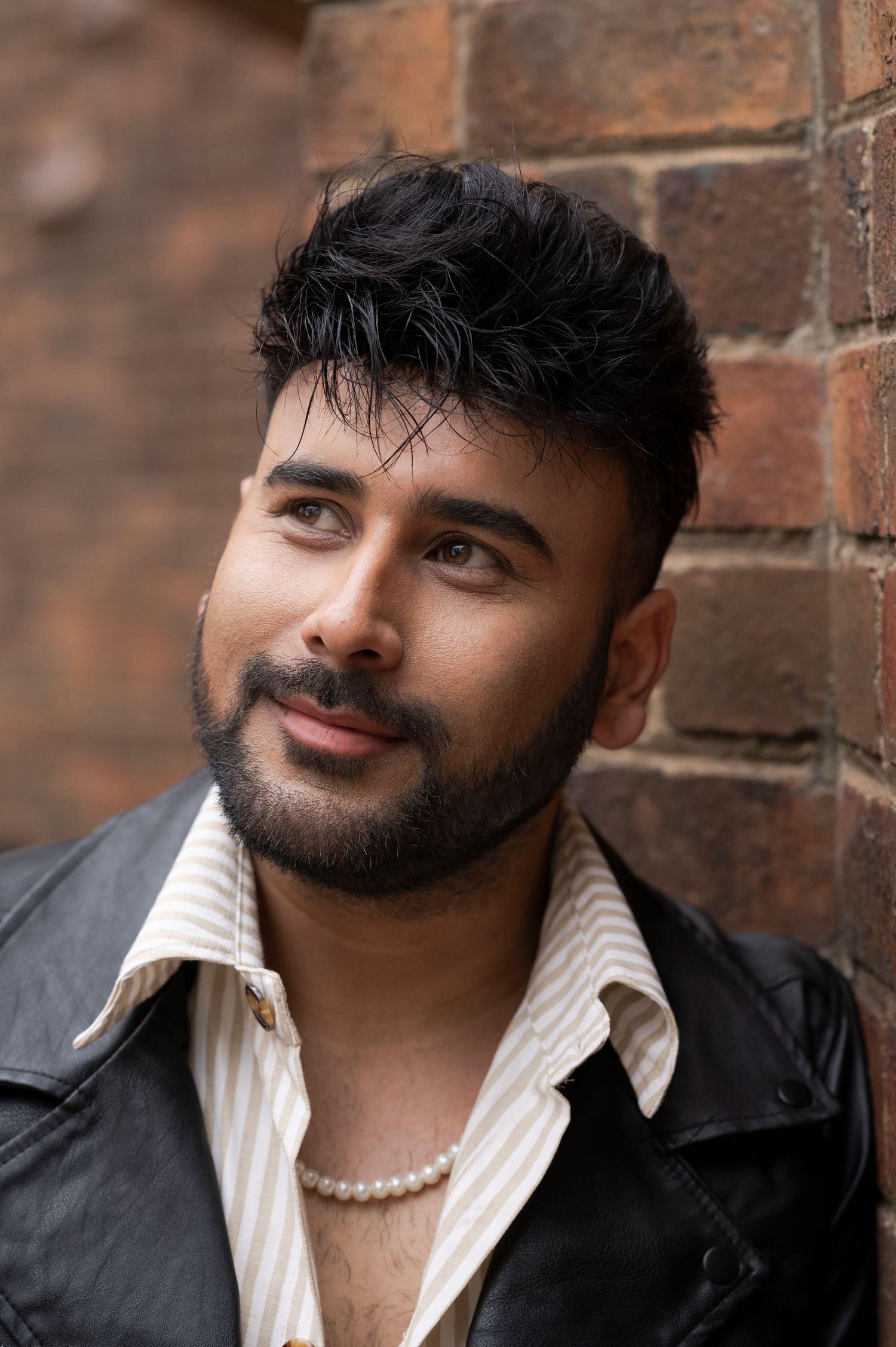 Young man with beard and pearl necklace leans against a brick wall wearing a leather jacket.